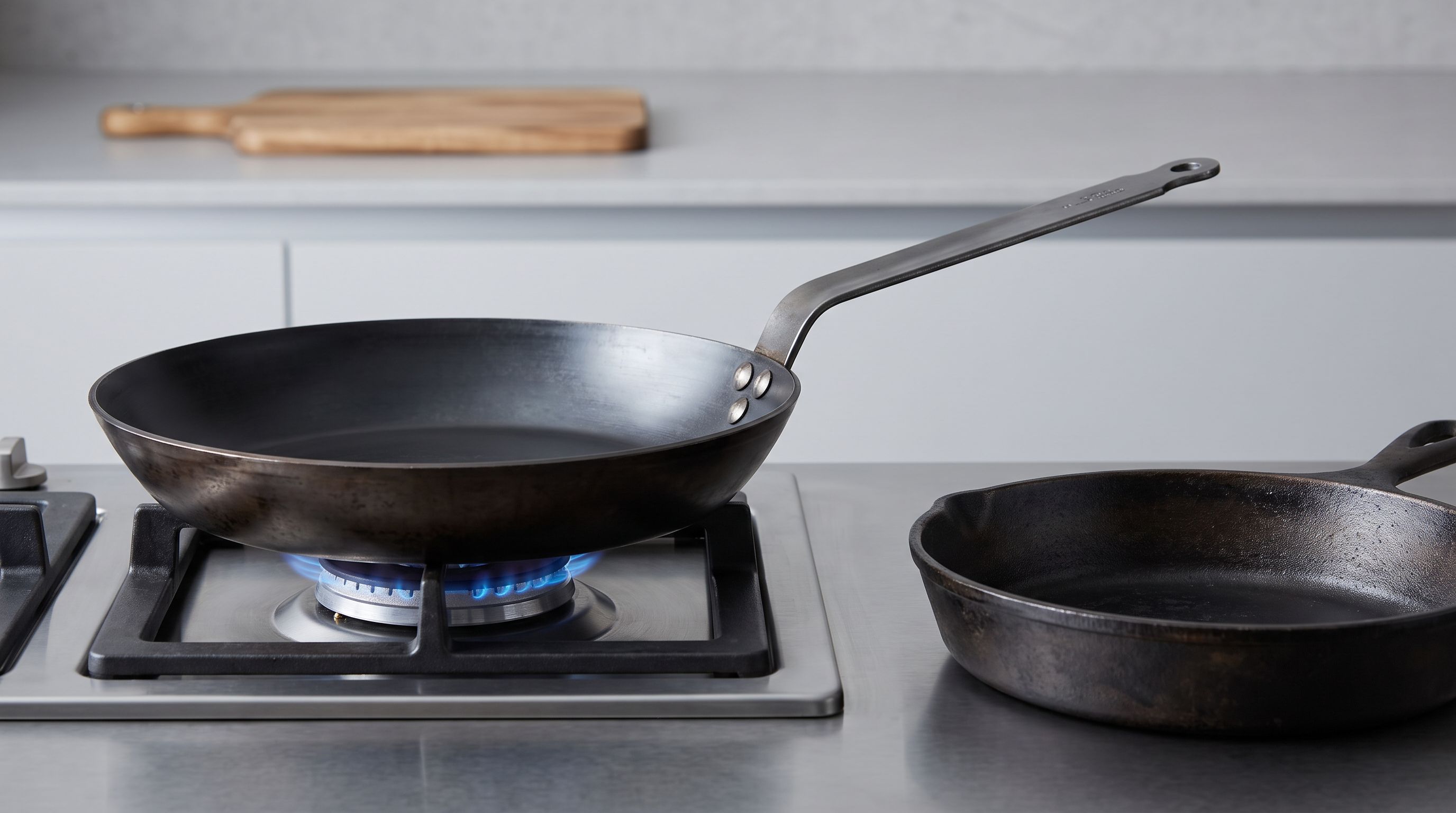 A dark, well-seasoned carbon steel skillet with a long riveted handle resting on a gas burner beside a smaller pre-seasoned pan