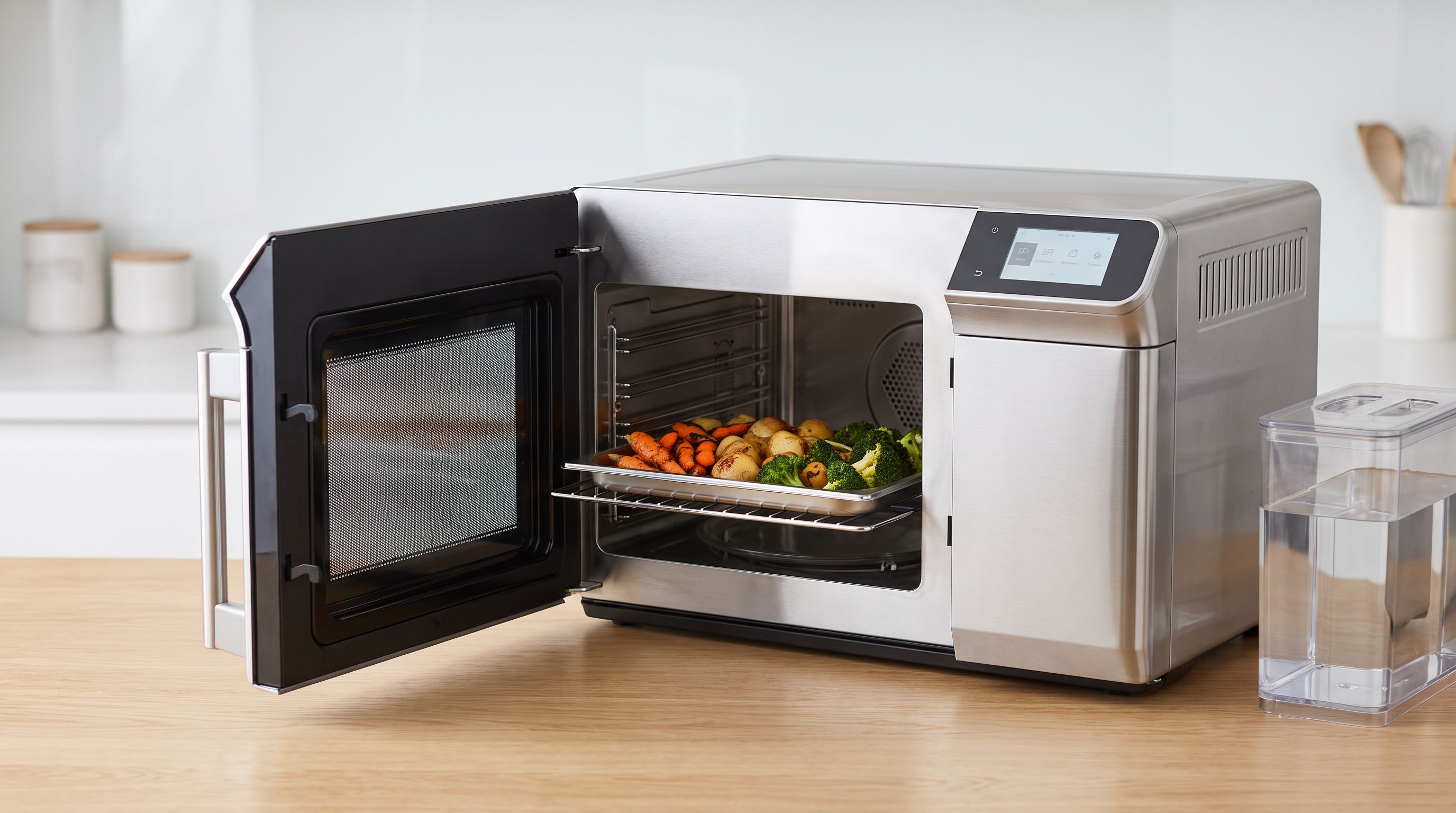 A stainless steel countertop combi steam oven on a kitchen counter with its glass door open, a water tank visible at the side and a rack of roasted vegetables inside