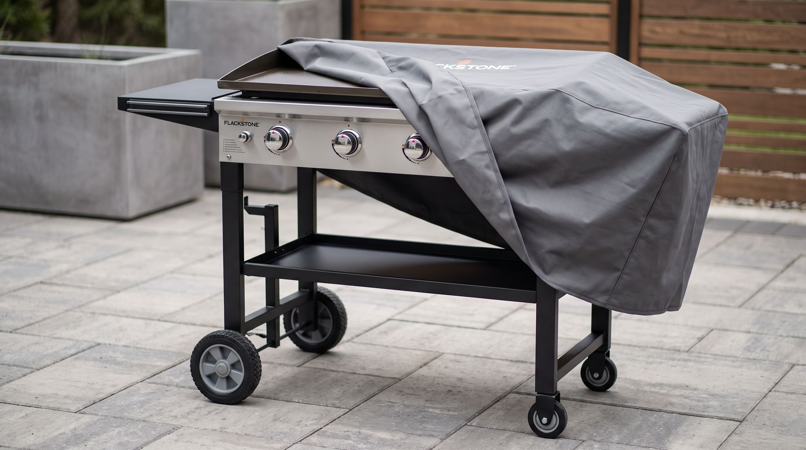 A 36-inch outdoor flat top griddle on a rolling cart with a heavy polyester cover partially pulled over the cooking surface on a patio