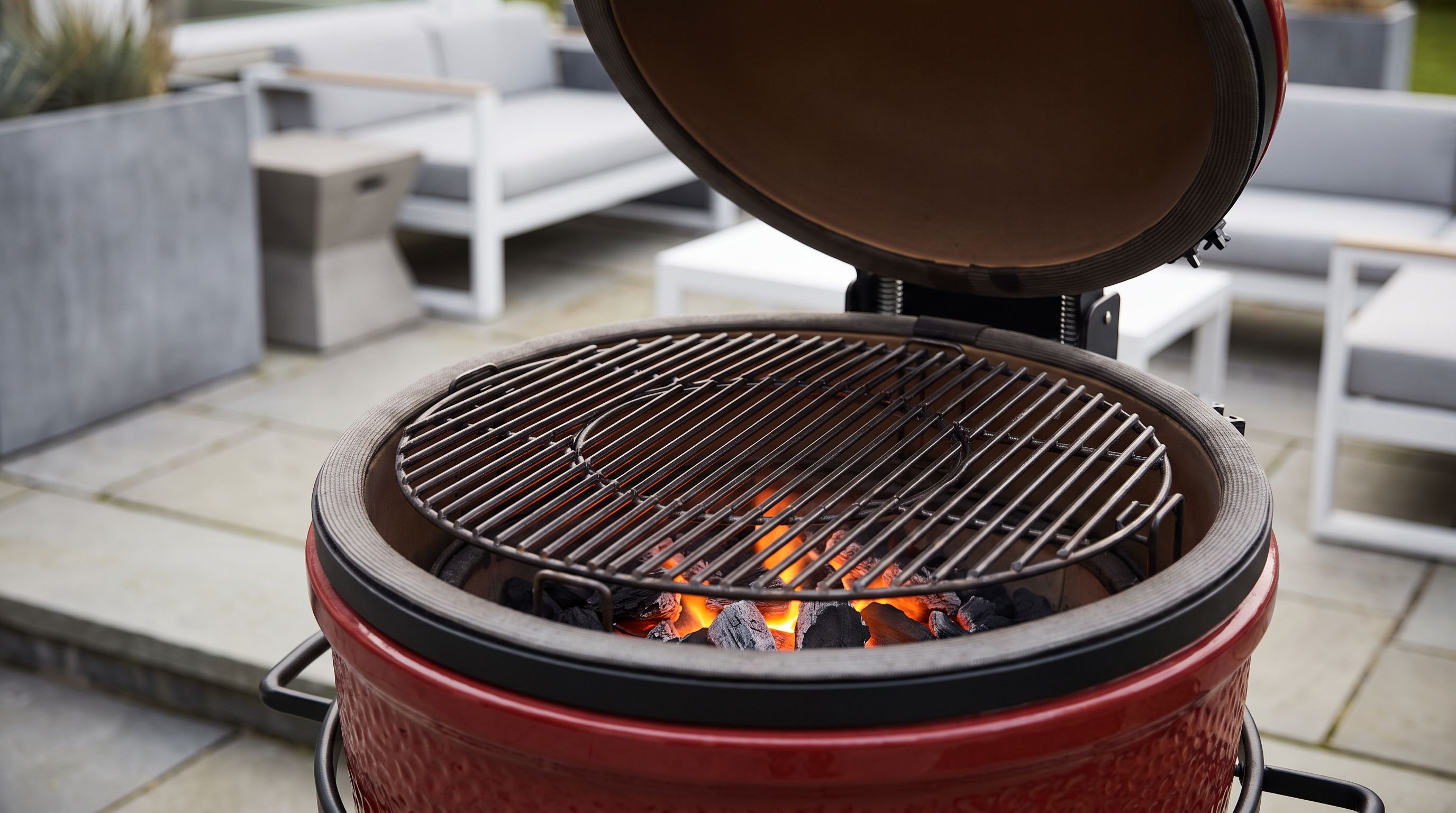A red ceramic kamado grill with its dome open on a patio, showing a round cast-iron cooking grate over glowing lump charcoal