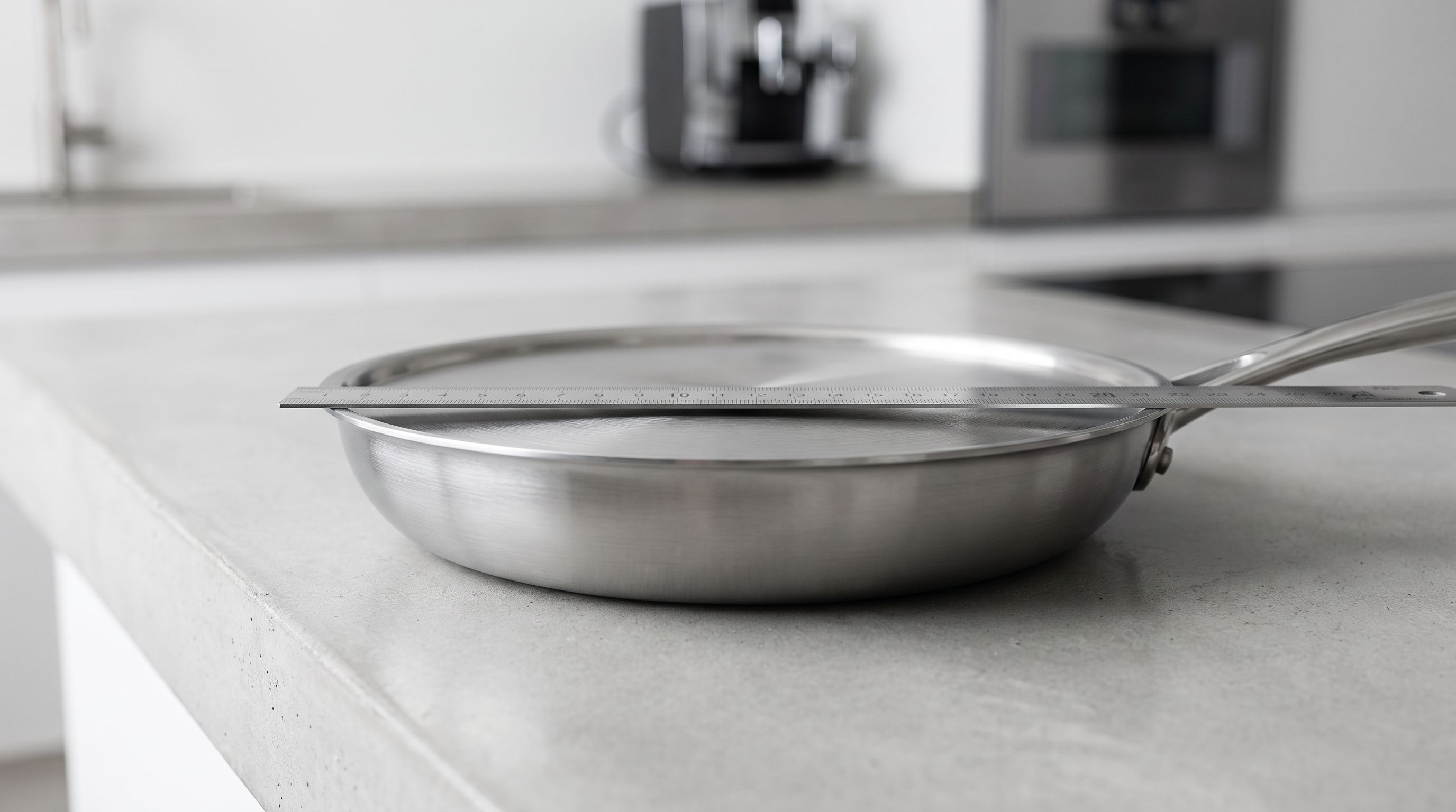 A stainless steel frying pan resting on a flat countertop with a straightedge laid across its base, showing a visible gap under the center