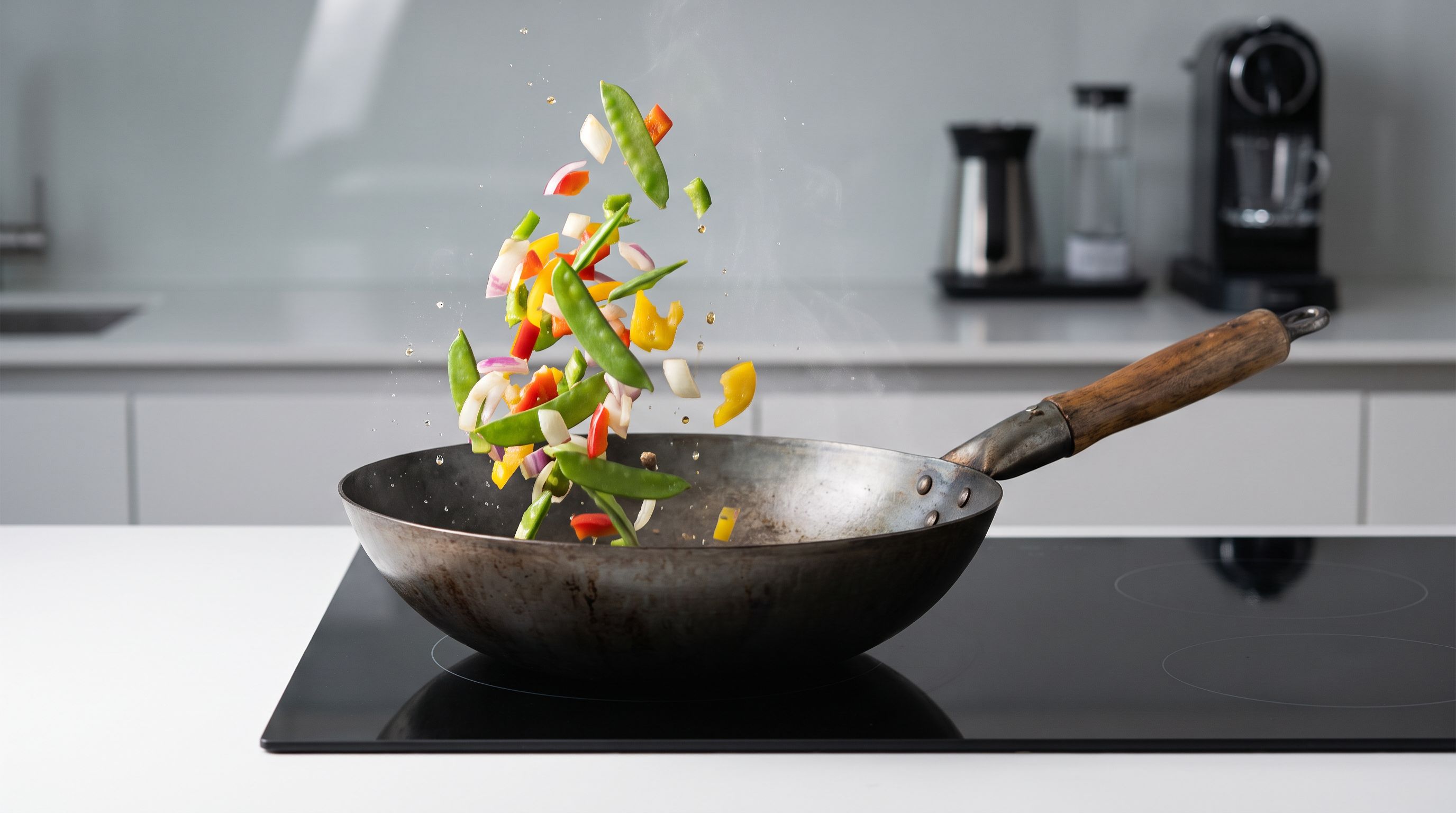 A seasoned flat-bottom carbon steel wok with a wooden handle sitting on an induction cooktop, vegetables mid-toss